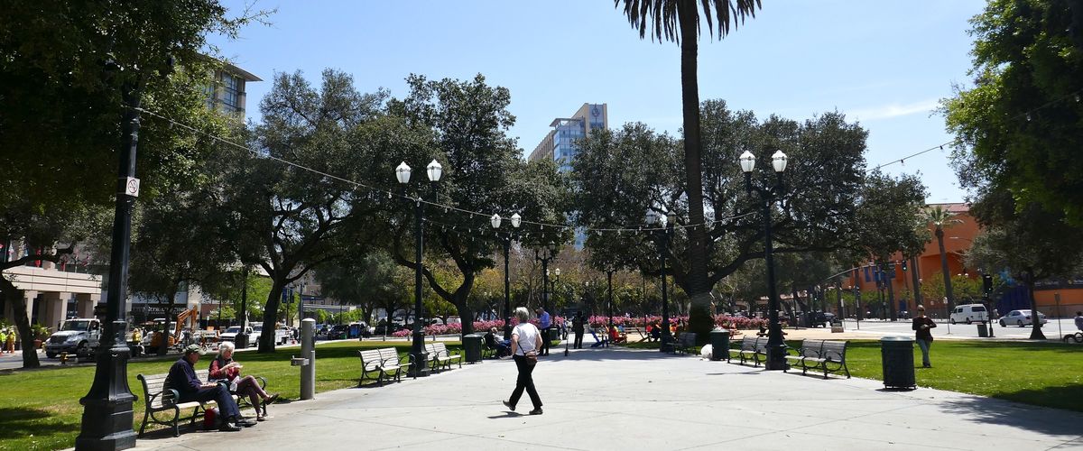 People walking and sitting on benches under palm and oak trees at Plaza de César Chávez, a free public park in Downtown San Jose